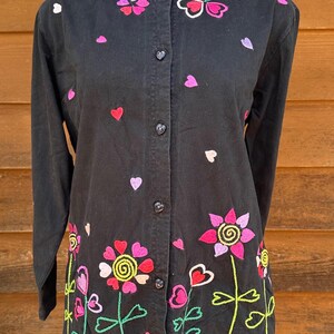 May include: Black button-up shirt with colorful embroidered flowers and hearts. The flowers have pink, green, yellow, and purple accents.