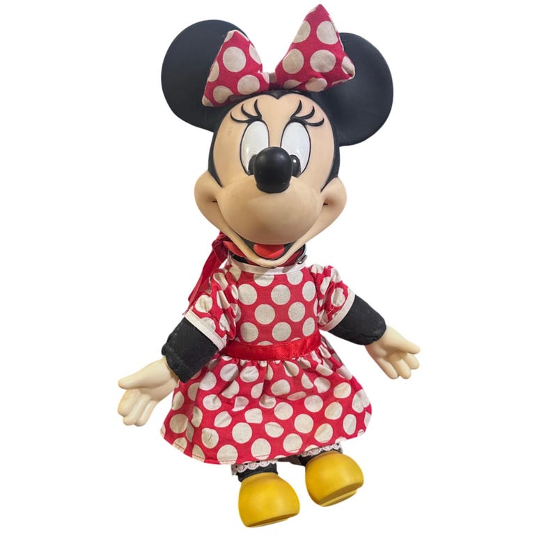 May include: A Minnie Mouse doll with a red and white polka dot dress, matching bow, and yellow shoes. The doll has black ears and a black nose. The doll's arms are outstretched.