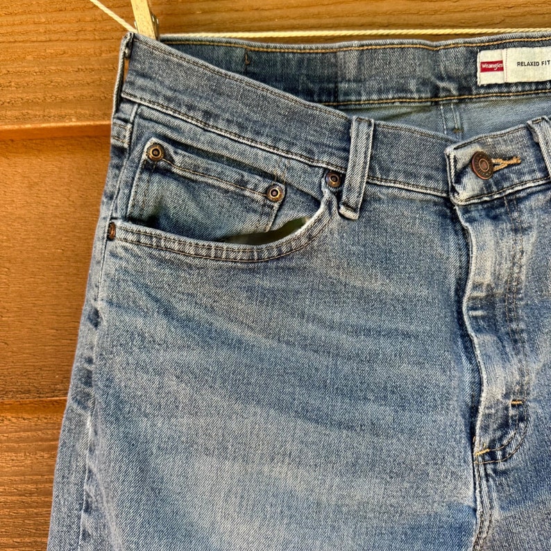 May include: A pair of blue denim jeans with a relaxed fit. The jeans have a front pocket and a button fly. The Wrangler brand label is visible on the right side of the waistband.