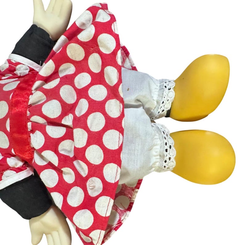 May include: A vintage doll with a red polka dot dress and white lace trim. The doll has yellow shoes and black sleeves. The doll's hands are visible, and the overall aesthetic is reminiscent of a classic toy.