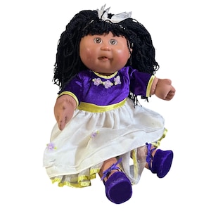 May include: A vintage doll with dark skin, black yarn hair, and brown eyes. The doll wears a purple velvet top with yellow trim, a white skirt, and purple velvet shoes. The doll's outfit is adorned with small floral accents.