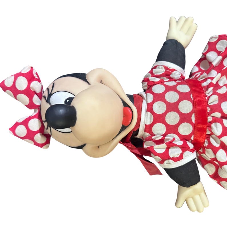 May include: A plush doll of a cartoon character with a large head, black eyes, and a black nose. The doll wears a red dress with white polka dots, a matching bow, and black sleeves. The doll's hands are visible.