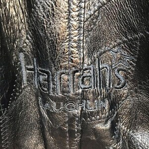 Può includere: Primo piano di una giacca in pelle nera con la scritta ricamata "Harrah's LAUGHLIN" in filo d'argento. La pelle ha un aspetto strutturato e rugoso con dettagli di cucitura visibili.