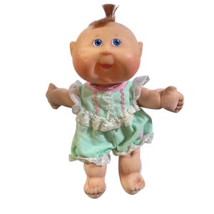 May include: A vintage Cabbage Patch Kid doll with a bald head and a tuft of brown hair. The doll has blue eyes and is wearing a mint green romper with white lace trim and a pink collar. The doll's arms are outstretched.