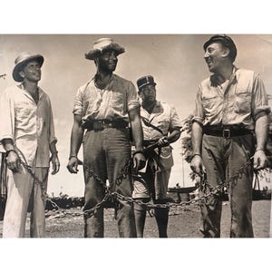 May include: Black and white photograph of four men. Three men are chained together, wearing light-colored shirts and pants, and straw hats. A man in a military uniform holds a rifle. The photograph appears to be taken outdoors.
