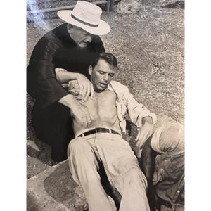 May include: Black and white photograph of two men. One man is wearing a white hat and a dark robe, and is kneeling. The other man is shirtless, wearing light-colored pants and a belt, and is lying down. The scene appears to be outdoors.