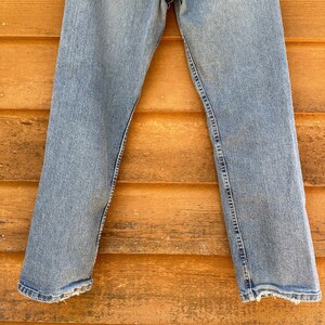 May include: A pair of light blue denim jeans with a straight leg fit. The jeans are hanging on a wooden wall.