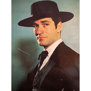 May include: A man in a black suit, vest, and a wide-brimmed black hat. The man is wearing a white collared shirt and a black bow tie. The background is a muted teal color.