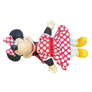 May include: A vintage Minnie Mouse plush doll, featuring a black head with large ears, a red and white polka dot bow, and a smiling face. She wears a red polka dot dress, white bloomers, black sleeves, and yellow shoes. The doll is lying down.
