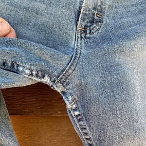 May include: Close-up of a pair of light blue denim jeans with a small tear in the fabric near the seam.