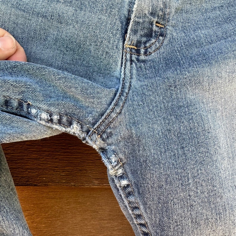 May include: Close-up of a pair of light blue denim jeans with a small tear in the fabric near the seam.