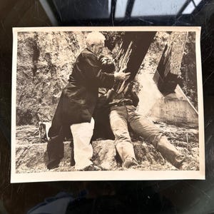 May include: Black and white photograph of two men interacting with a large wooden structure. One man, wearing a long coat, appears to be assisting the other, who is wearing jeans. The background features a rocky terrain.