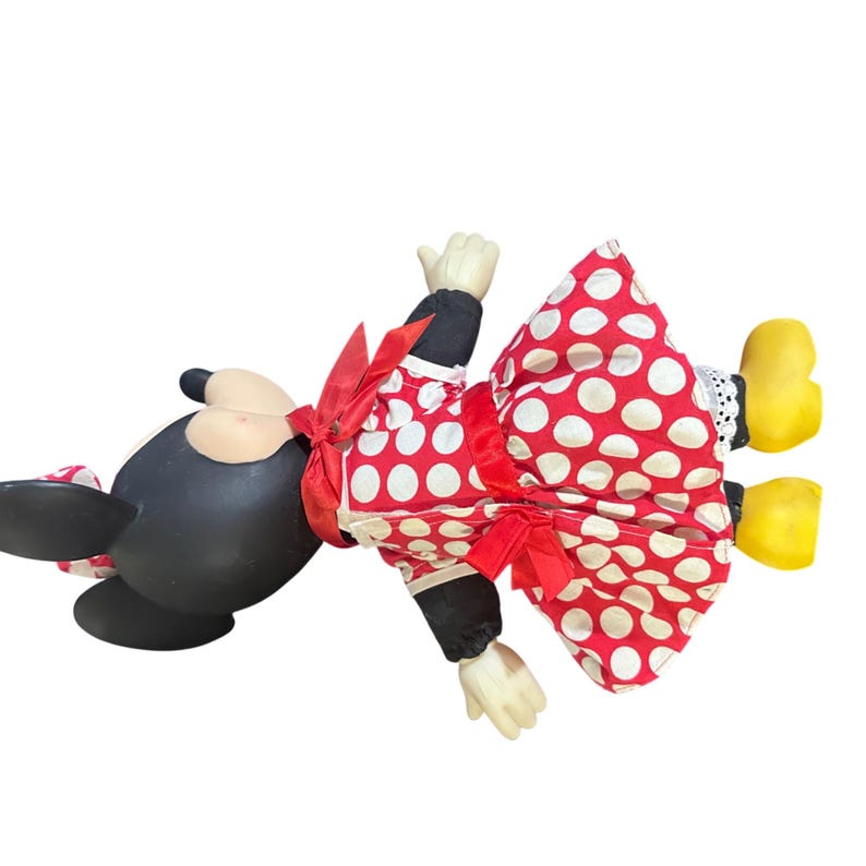 May include: A Minnie Mouse doll, lying on its side, features a black head and body, with a red and white polka dot dress. The doll has white gloves, yellow shoes, and a red bow. The dress has a red ribbon around the waist.