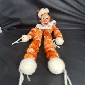 May include: A crocheted clown doll with orange and white stripes. The clown has a white hat and a painted face with a red smile.