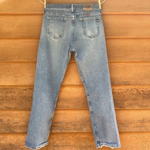 May include: A pair of light blue denim jeans with a faded look. The jeans have a straight leg and a frayed hem. The Wrangler logo is visible on the back pocket.