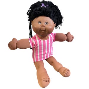 Könnte beinhalten: Eine Vintage Cabbage Patch Kid-Puppe mit dunkler Haut, schwarzen Garnhaaren und einem rosa-weiß gestreiften Hemd. Die Puppe hat braune Augen und ist mit ausgestreckten Armen posiert. Eine lila Schleife akzentuiert das Haar.