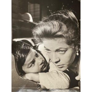 May include: A black and white photograph of a woman looking down at a young boy. The woman has her hair styled in a bouffant and is wearing a pearl earring. The boy is looking up at the woman with a serious expression.