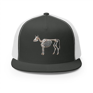 May include: A black and white trucker cap with a cow skeleton design. The front panel is black with a detailed illustration of the skeletal structure. The mesh back is white, and the brim is black.