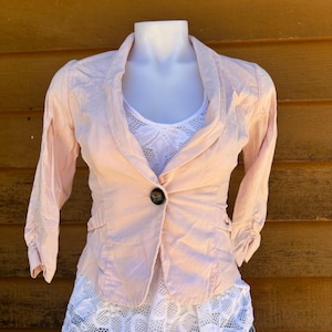 Puede incluir: Una chaqueta de color rosa pálido con un cierre de botón único y mangas largas. La chaqueta está hecha de una tela ligera y tiene un ajuste holgado.