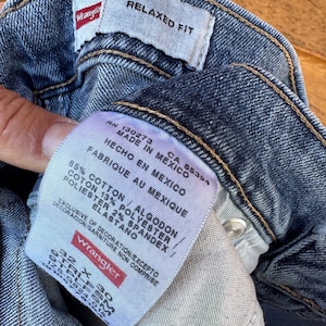 May include: A blue denim jean label with white text that reads "Relaxed Fit", "RN 130273 CA 55394", "Made in Mexico", "Hecho en Mexico", "Fabrique au Mexique", "85% Cotton / Algodon", "Coton 13% Polyester", "Polyester 2% Spandex / Elastano", "Exclusive of decoration/Excepto decoracion/Garniture non comprise", and "Wrangler".