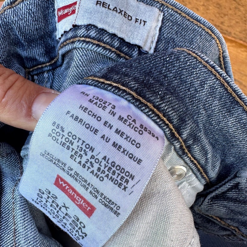 May include: A blue denim jean label with white text that reads "Relaxed Fit", "RN 130273 CA 55394", "Made in Mexico", "Hecho en Mexico", "Fabrique au Mexique", "85% Cotton / Algodon", "Coton 13% Polyester", "Polyester 2% Spandex / Elastano", "Exclusive of decoration/Excepto decoracion/Garniture non comprise", and "Wrangler".