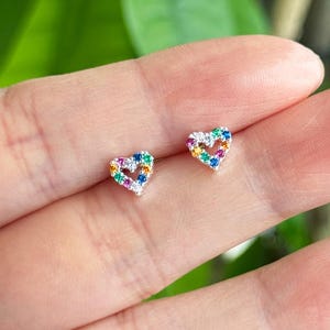 Könnte beinhalten: Ein Paar herzförmige Ohrstecker. Jeder Ohrring ist mit bunten Edelsteinen besetzt, wodurch ein Regenbogeneffekt entsteht. Die Ohrringe sind klein und zart, perfekt für den täglichen Gebrauch. Die Ohrringe sind aus Silber oder weißem Metall gefertigt.