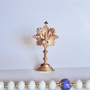 Puede incluir: Una custodia religiosa dorada con una cruz en la parte superior y una paloma en el centro. La custodia está sobre una superficie blanca, con un rosario de cuentas blancas y doradas en primer plano.