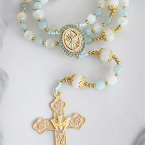 Puede incluir: Un rosario con cuentas celestes y blancas, acentuado con metal dorado. La cruz al final es rosa con detalles dorados y un emblema de paloma. El rosario presenta una pieza central ovalada con un dise&ntilde;o de paloma.