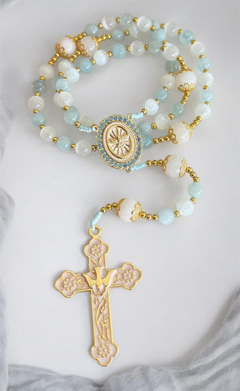Puede incluir: Un rosario con cuentas celestes y blancas, acentuado con metal dorado. La cruz al final es rosa con detalles dorados y un emblema de paloma. El rosario presenta una pieza central ovalada con un dise&ntilde;o de paloma.