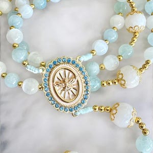 Puede incluir: Un rosario con cuentas de piedras preciosas azul claro y blancas, con detalles dorados. La pieza central es un colgante ovalado con un dise&ntilde;o dorado y un borde de cristal azul. El rosario tiene una apariencia delicada y ornamentada.