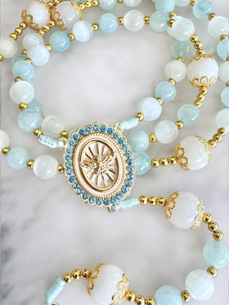 Puede incluir: Un rosario con cuentas de piedras preciosas azul claro y blancas, con detalles dorados. La pieza central es un colgante ovalado con un dise&ntilde;o dorado y un borde de cristal azul. El rosario tiene una apariencia delicada y ornamentada.