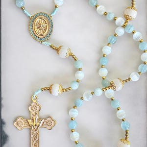 Puede incluir: Un rosario con cuentas azul claro y blancas, detalles dorados y un colgante de cruz dorada. La cruz presenta un dise&ntilde;o detallado con una paloma en el centro. Las cuentas blancas decorativas est&aacute;n intercaladas.