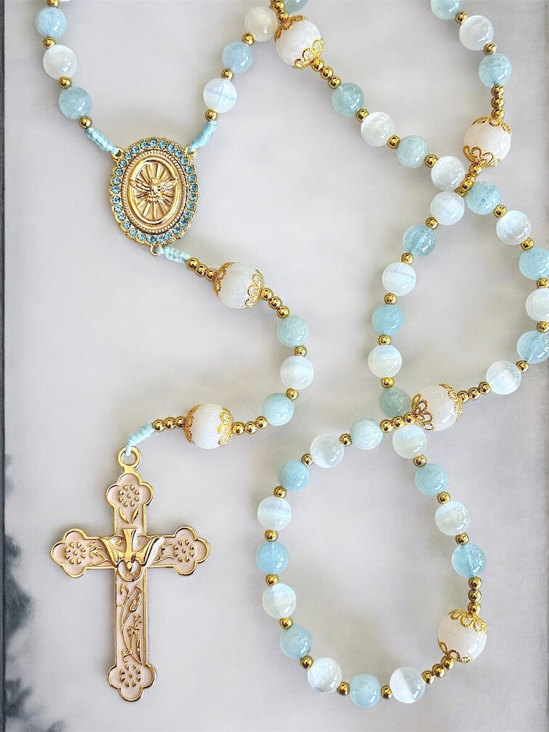 Puede incluir: Un rosario con cuentas azul claro y blancas, detalles dorados y un colgante de cruz dorada. La cruz presenta un dise&ntilde;o detallado con una paloma en el centro. Las cuentas blancas decorativas est&aacute;n intercaladas.