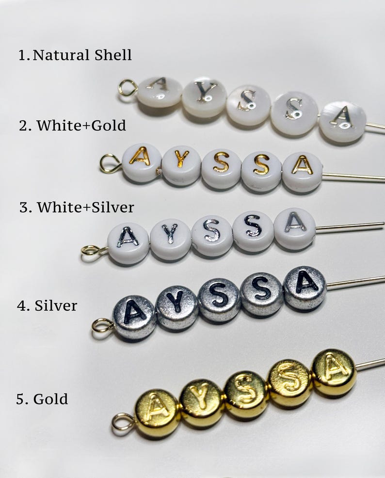 Puede incluir: Primer plano de cinco estilos diferentes de cuentas que deletrean el nombre "AYSSA". Las cuentas son blancas, plateadas, doradas y de concha natural, con las letras en varios acabados met&aacute;licos. Cada hebra est&aacute; en un alfiler plateado.