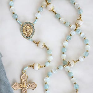 Puede incluir: Un rosario con cuentas azul claro y blancas, detalles dorados y un colgante de cruz dorada. Las cuentas est&aacute;n intercaladas con cuentas blancas m&aacute;s grandes y conectadas por metal dorado. La cruz tiene un acabado dorado rosado.