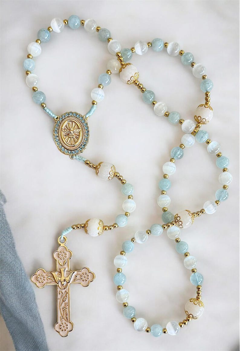 Puede incluir: Un rosario con cuentas azul claro y blancas, detalles dorados y un colgante de cruz dorada. Las cuentas est&aacute;n intercaladas con cuentas blancas m&aacute;s grandes y conectadas por metal dorado. La cruz tiene un acabado dorado rosado.