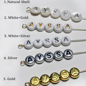 Puede incluir: Cinco filas de cuentas redondas en alfileres de metal, cada una deletreando A Y S S A. Fila 1: concha natural con letras plateadas. Fila 2: cuentas blancas con letras doradas. Fila 3: cuentas blancas con letras plateadas. Fila 4: cuentas plateadas.