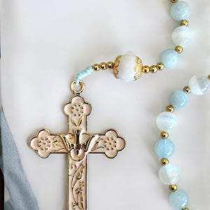 Puede incluir: Un rosario con una cruz dorada y cuentas. La cruz es de color rosa p&aacute;lido con detalles dorados y un dise&ntilde;o de paloma. Las cuentas son una mezcla de azul claro y blanco, separadas por espaciadores dorados. El rosario est&aacute; sobre un fondo blanco.