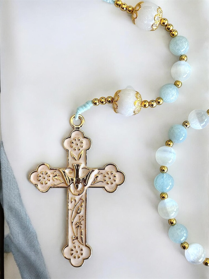 Puede incluir: Un rosario con una cruz dorada y cuentas. La cruz es de color rosa p&aacute;lido con detalles dorados y un dise&ntilde;o de paloma. Las cuentas son una mezcla de azul claro y blanco, separadas por espaciadores dorados. El rosario est&aacute; sobre un fondo blanco.