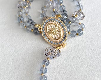 Puede incluir: Un rosario con cuentas azul-grisáceas y detalles dorados. La pieza central es un colgante ovalado con un diseño dorado y un borde de piedras preciosas azules. También hay dos cuentas claras en forma de rosa.