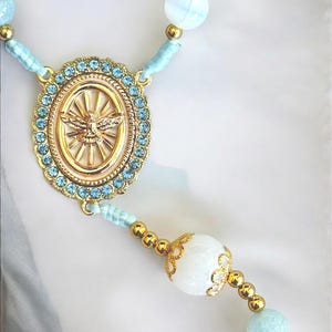 Puede incluir: Un rosario con una pieza central dorada con un emblema de paloma, rodeada de cristales azul claro. El rosario tiene cuentas azules claras y blancas con detalles dorados, creando una est&eacute;tica delicada y espiritual.