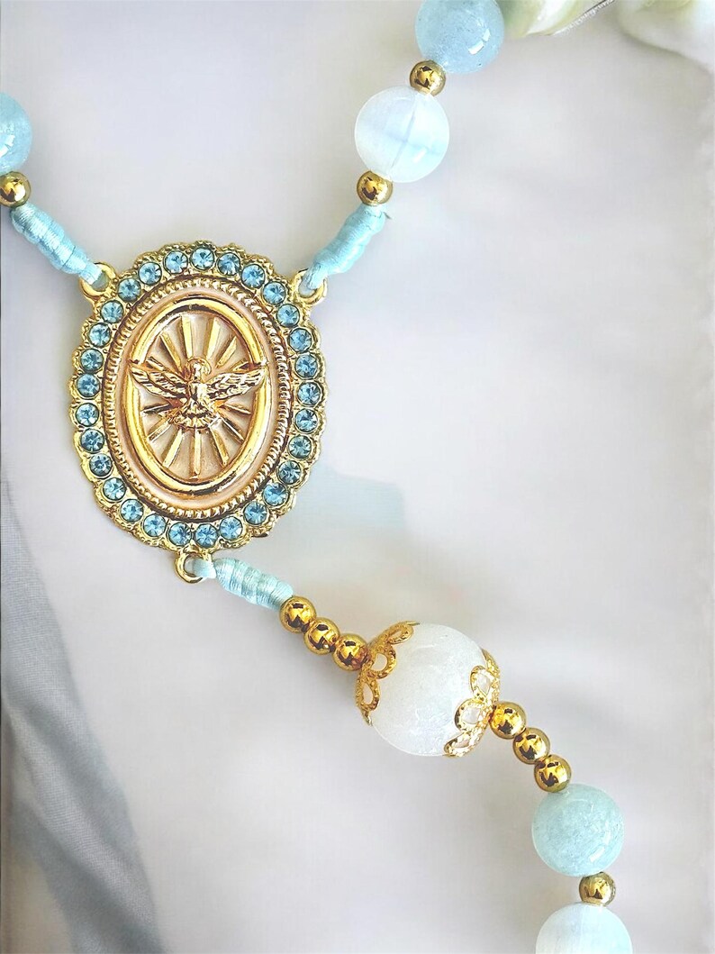 Puede incluir: Un rosario con una pieza central dorada con un emblema de paloma, rodeada de cristales azul claro. El rosario tiene cuentas azules claras y blancas con detalles dorados, creando una est&eacute;tica delicada y espiritual.