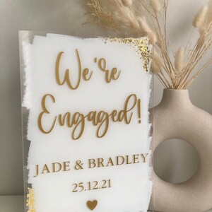 May include: White acrylic sign with gold lettering announcing "We're Engaged!" with the names Jade & Bradley and the date 25.12.21. The sign has a gold glitter effect around the edges.
