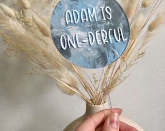 May include: A blue and white marbled cake topper with the text "Adam is one-derful" in white lettering.