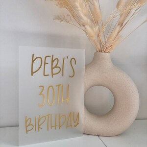 May include: A frosted acrylic sign with gold lettering that reads "DEBI'S 30TH BIRTHDAY".