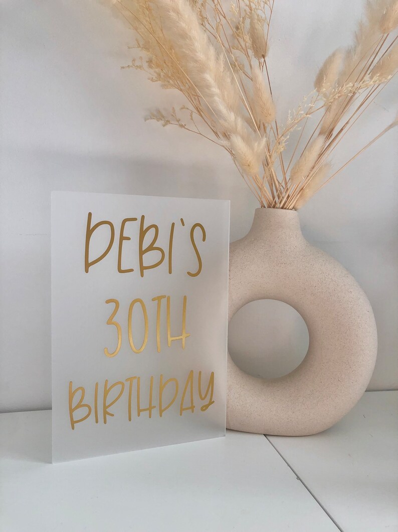 May include: A frosted acrylic sign with gold lettering that reads "DEBI'S 30TH BIRTHDAY".