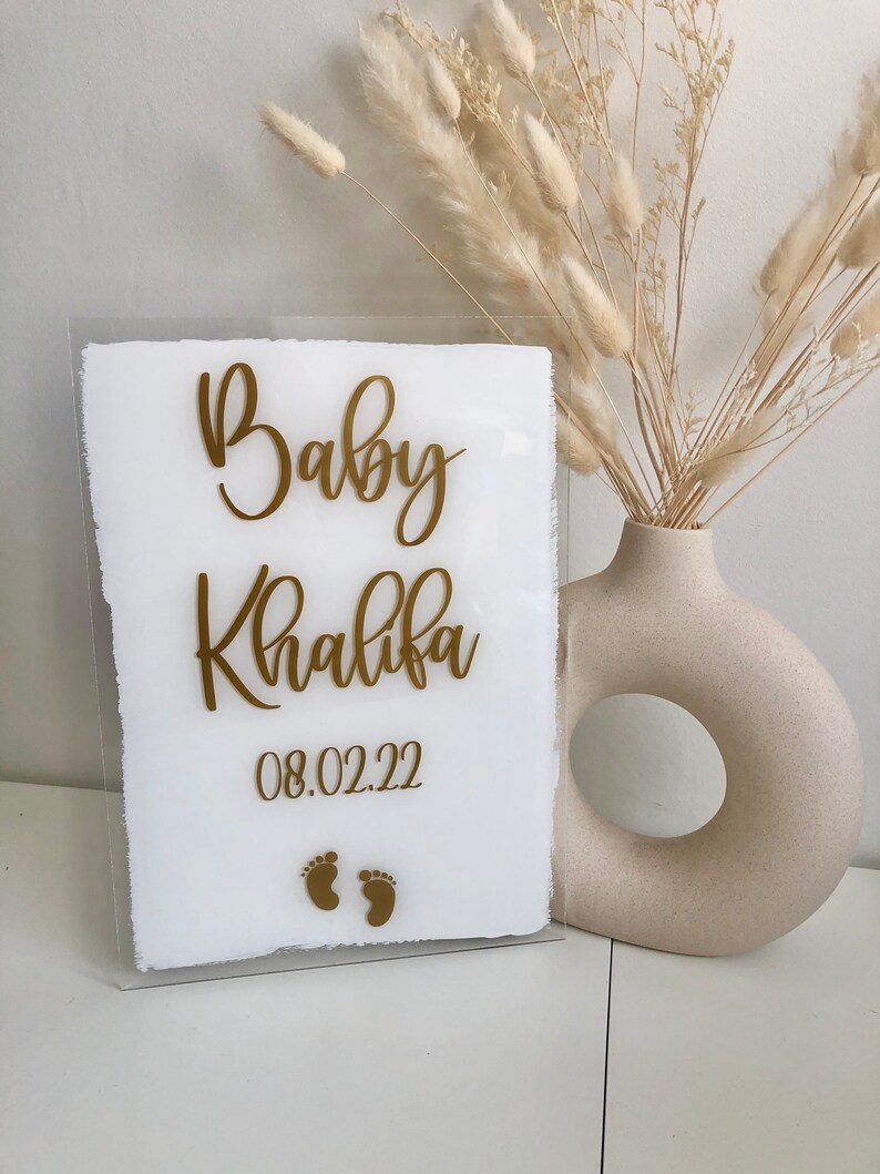 May include: A white acrylic sign with gold lettering that reads "Baby Khalifa 08.02.22" with two gold footprints below the text.