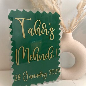 May include: A clear acrylic sign with a green painted background and gold lettering. The sign reads "Tahir's Mehndi! 28 January 2022".