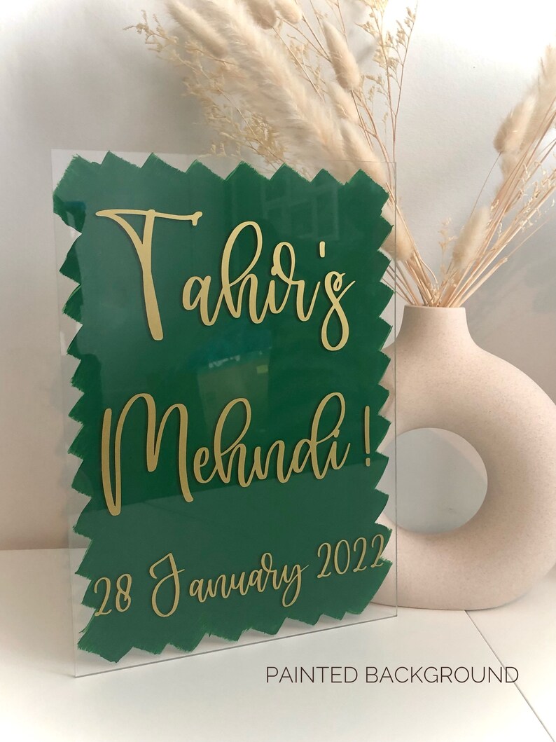 May include: A clear acrylic sign with a green painted background and gold lettering. The sign reads "Tahir's Mehndi! 28 January 2022".