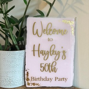 May include: A pink and gold welcome sign with the text "Welcome to Hayley's 50th Birthday Party".